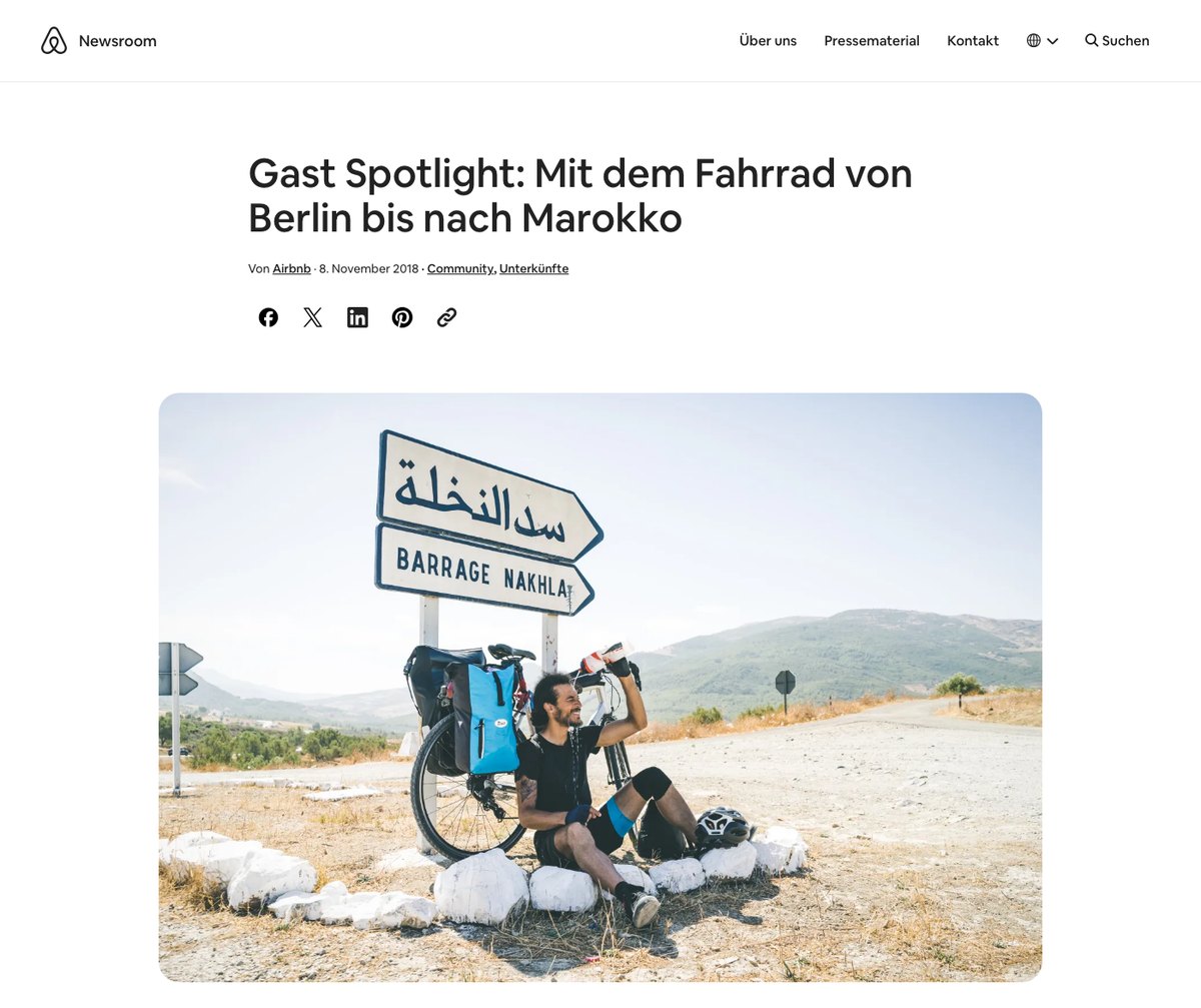 Screenshot of the Airbnb Newsroom article “Gast Spotlight: Mit dem Fahrrad von Berlin bis nach Marokko”, showing Ahmed resting beside a loaded touring bicycle under a road sign in Morocco.